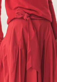 Roter plissierter Rock mit einer gebundenen Schleifen-Detail an der Taille. Aus strukturiertem Stoff gefertigt, der eine glatte Textur und eine lebendige Farbe aufweist.