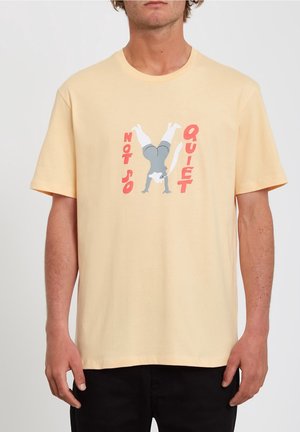 Mann trägt ein hellgelbes T-Shirt mit einem Motiv eines grauen Katers, der einen Handstand macht, und dem Text "NICHT SO LEISE" in roten Buchstaben.