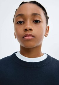 Detailní záběr na mladou osobu s copánky, oblečenou v tmavě modrém svetru přes bílou košili, hledící přímo vpřed s neutrálním výrazem.