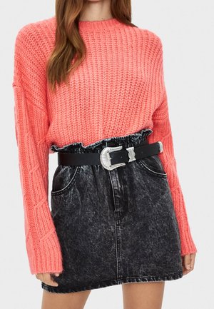 Vrouw draagt een koraalkleurige, grofgebreide trui die is ingestopt in een zwarte acid-wash denim minirok met een decoratieve zilveren gesp.