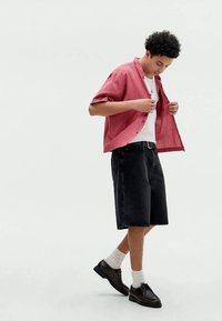 Camisa de mangas curtas, xadrez vermelho sobre uma t-shirt branca, combinada com calções de ganga pretos e sapatos pretos, com meias brancas.