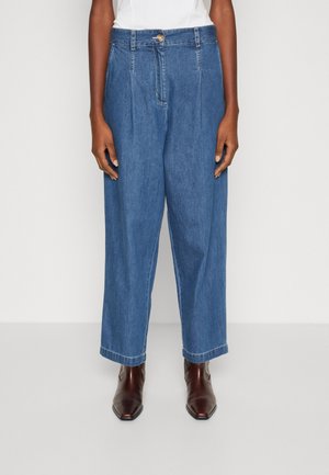 Personne portant un pantalon en jean bleu taille haute, coupe ample, associé à des bottes pointues marron foncé, debout devant un fond clair uni.