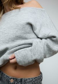 Pull en tricot gris clair, décolleté sur les épaules, avec des poignets à côtes. Texture douce, design court révélant le ventre, associé à un jean gris déchiré.