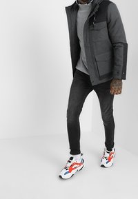 Grå hættejakke med en sort yoke, fire lommer, båret over en lys grå skjorte og sorte jeans. Farverige sneakers med røde, blå og hvide accenter.