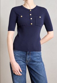 Granatowy prążkowany top z krótkimi rękawami, z okrągłym dekoltem, pięcioma guzikami w kolorze złotym oraz dwoma kieszeniami na piersi. Noszony z niebieskimi dżinsami.