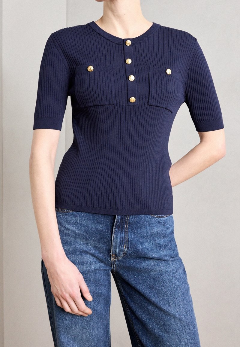 Granatowy prążkowany top z krótkimi rękawami, z okrągłym dekoltem, pięcioma guzikami w kolorze złotym oraz dwoma kieszeniami na piersi. Noszony z niebieskimi dżinsami.