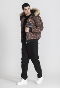Giacca a vento marrone con motivo e cappuccio foderato in pelliccia, chiusura con zip e tasche frontali, abbinata a pantaloni neri e sneakers bianche.