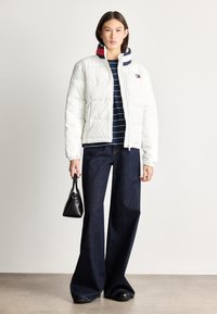 Balta pūkinė striukė su siuvinėtu raštu, papuošta daugiakampiu juostuotu kapišonu. Derinama su tamsiomis plačiomis kelnėmis ir mažais juodais rankinukais.