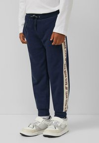Navyblaue Sweatpants aus Baumwolle mit einem gerippten Bund, seitlichen Streifen mit dem Text "TEAM ROAR" und Bündchen, kombiniert mit weißen Sneakers.