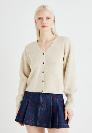 Beige ribbestrikket cardigan med V-hals, som har seks brune knapper. Kombinert med en blå denimskjørt prydet med røde sommerfugler broderi.