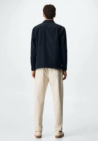 Ciemnoniebieska koszula z długim rękawem o prostym kroju, w połączeniu z jasnobeżowymi spodniami. Model stoi tyłem, eksponując teksturę tkaniny i dopasowanie.