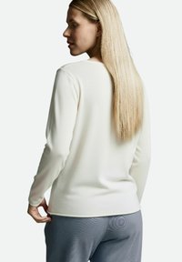 Weißes langärmeliges Oberteil mit strukturierter Oberfläche, rundem Ausschnitt, figurbetontem Schnitt und leicht verlängertem Saum, kombiniert mit grauer Hose.