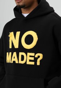 Zwarte hoodie met grote gele geborduurde tekst "NO MADE?" op de voorkant. Het materiaal lijkt zacht met een ontspannen pasvorm en een kangoeroezak.