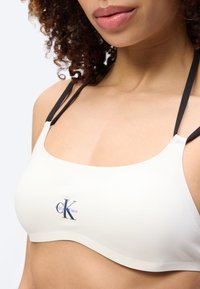 Γυναίκα που φοράει λευκό αθλητικό σουτιέν Calvin Klein με μαύρα λουριά, εμφανίζοντας τον λαιμό, τους ώμους και μερικό πρόσωπο με σγουρά μαλλιά.