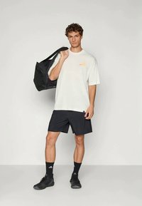 Hvit oversized t-skjorte med liten oransje trykk, svarte treningsshorts, svarte sko og en svart treningssekk. Enkelt, avslappet sportstøy-uttrykk.
