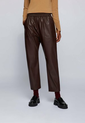 Person trägt dunkelbraune Kunstlederhose mit elastischem Bund, beigen Pullover, bordeauxrote Socken und schwarze Loafers und steht vor einfarbigem Hintergrund.