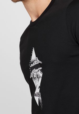 Mężczyzna noszący czarną koszulkę z białą grafiką stylizowanych gór i chmur oraz tekstem "THE NORTH FACE" ustawionym pionowo.