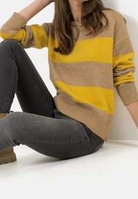 Strikket sweater i beige og gule striber, med rund hals og ribbede ærmer. Sammen med mørkegrå jeans og brune støvler, siddende mod en hvid baggrund.