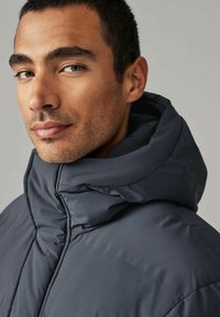 Dunkelgraue Daunenjacke mit hohem Kragen und einer großen, gefütterten Kapuze. Glatte Textur mit sichtbaren Nähdetails und einer taillierten Passform.
