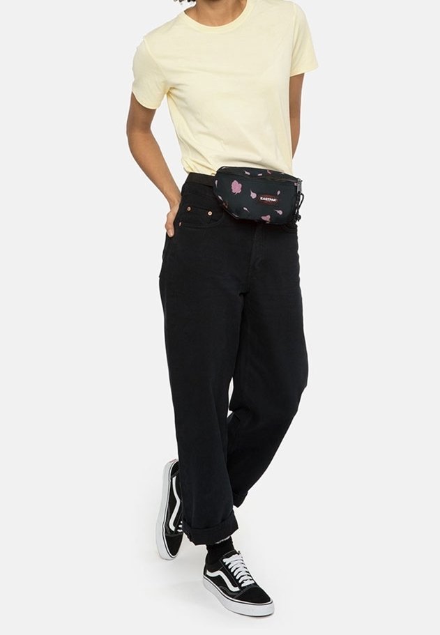 Svart midjeväska med blommönster, bärs över en blekgul T-shirt och svarta vida byxor. Svart och vita sneakers kompletterar outfiten.