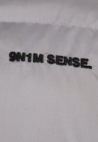 Sølvstof med en glat overflade præsenterer fed sort broderet tekst, der læser "9N1M SENSE." Ingen yderligere mønstre eller hardware synlig.