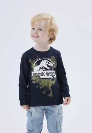 Camisa de manga larga de color azul marino con gráfico de dinosaurio y texto "Jurassic World"; tejido suave, cuello redondo y ajuste relajado. Pantalones cargo de mezclilla claros.