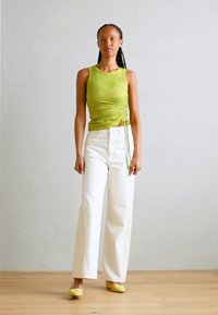 Top a righe verde con lati arricciati, abbinato a pantaloni bianchi a vita alta e scarpe piatte gialle. Pavimento in legno, sfondo neutro.
