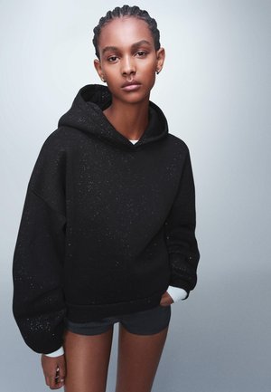 Čierna oversize mikina s kapucňou, zdobená škvrnitým vzorom, so šnúrkou na kapucni a kontrastnými bielymi manžetami. Model má na sebe tmavé šortky.
