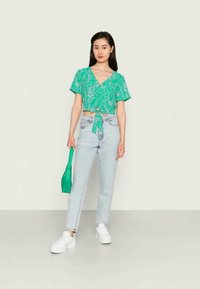 Top curto verde com mangas curtas e padrão floral, amarrado na cintura, combinado com jeans de perna reta de lavagem clara e ténis brancos.