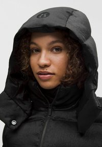 Chaqueta acolchada negra con capucha, que cuenta con un cierre de cremallera y un logo en la capucha. La tela tiene una textura suave y un acabado brillante.