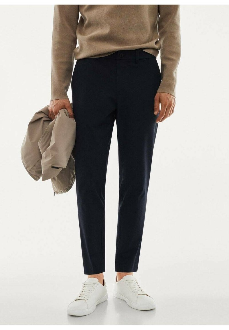 Navy slim-fit-housut, jotka on valmistettu sileästä kankaasta, yhdistettynä beigeen neuleeseen ja vaaleaan takkiin. Asusteena valkoiset lenkkarit.