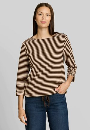 Vrouw met stijl bruin haar die een bruin-beige gestreepte top met lange mouwen en een blauwe spijkerbroek draagt, staand tegen een effen grijze achtergrond.