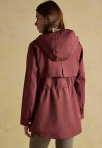 Bordowa kurtka z kapturem i ściągniętym w pasie, z detalem wentylacyjnym z tyłu i długimi rękawami. Materiał wydaje się gładki i wodoodporny. Widoczne kraciaste spodnie.