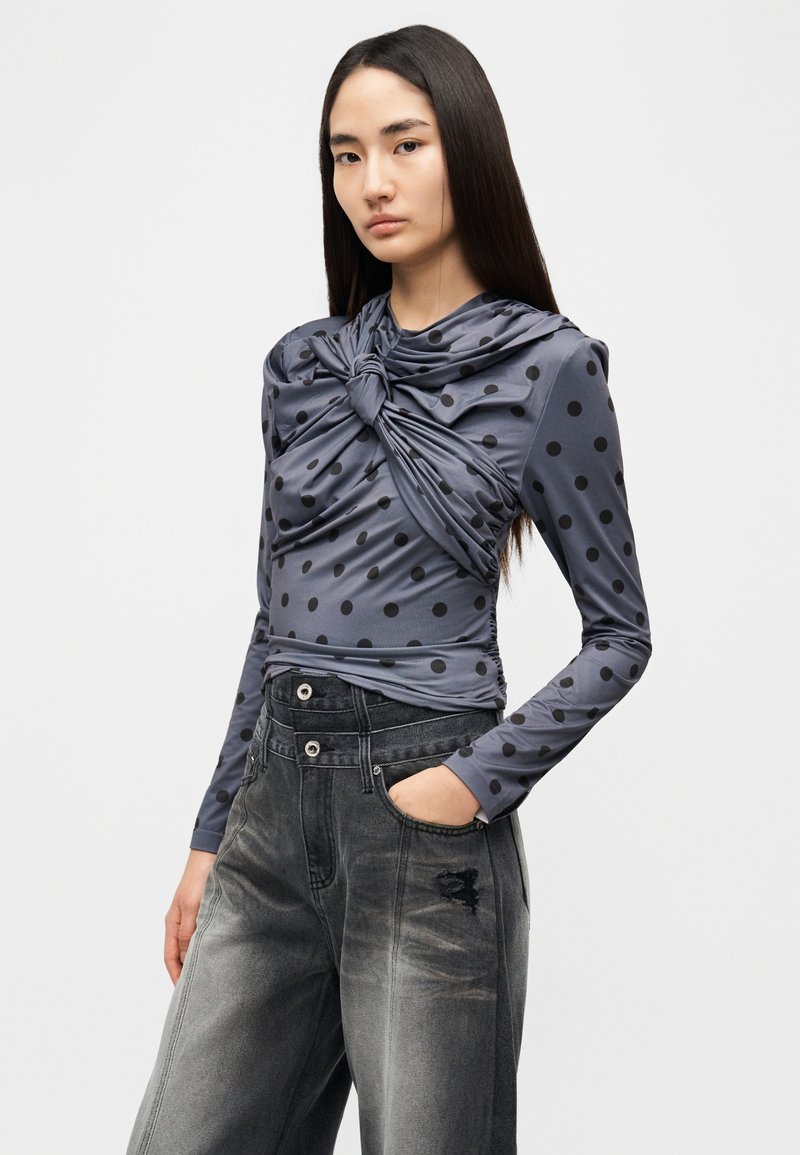 Haut à manches longues gris avec des pois noirs, doté d'un détail torsadé au niveau de l'encolure. Associé à un jean en denim usé et taille haute.