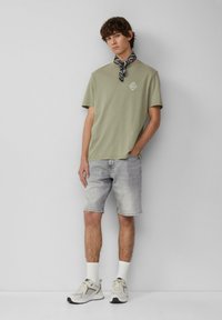 Oliwkowa bawełniana koszulka z małym logo, zestawiona z wyblakłymi szarymi szortami dżinsowymi. Białe skarpetki do kostek i białe sneakersy z różnych materiałów.