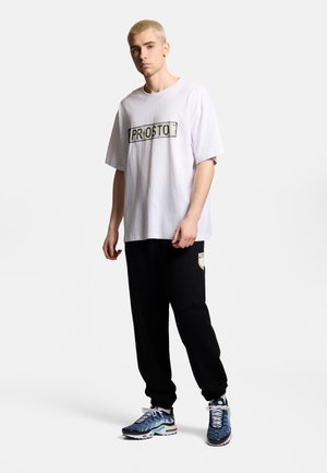 Młody mężczyzna z krótkimi blond włosami, ubrany w białą koszulkę "PROSTO", czarne spodnie dresowe i niebieskie buty sportowe, stojący na białym tle.