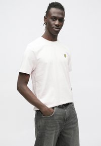 Lichtroze katoenen T-shirt met een ronde hals en korte mouwen, met een klein geel logo op de linkerbuste. Draagt dit met grijze spijkerbroek.