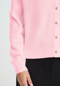 Rosa stickad cardigan med ribbade ärmslut och nederkant. Har dekorativa knappar framtill som visar upp ett texturerat tyg. Svarta byxor är synliga.