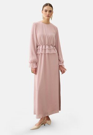 Kvinde iført en lang lyserød kjole med lagdelt flæsetalje og lange ærmer, kombineret med beige spidse lavhælede sko og guldbelagte øreringe.