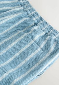 Pantalon en tissu texturé rayé bleu et blanc avec une ceinture élastique et une grande poche avant, étalé à plat sur une surface blanche.