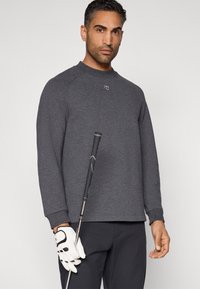 Σκούρο γκρι πουλόβερ με στρογγυλή λαιμόκοψη, ριμπ μανσέτες και λογότυπο στο στήθος. Το μοντέλο κρατάει ένα γκολφ κλαμπ, φοράει λευκό γάντι γκολφ.