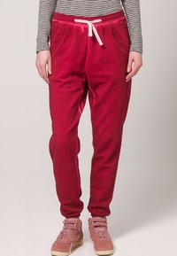 Sarkani sporta bikses ar elastīgu jostu un auklu, kurām ir sānu kabatas un brīva piegriezuma, papildinātas ar rozā kedām.