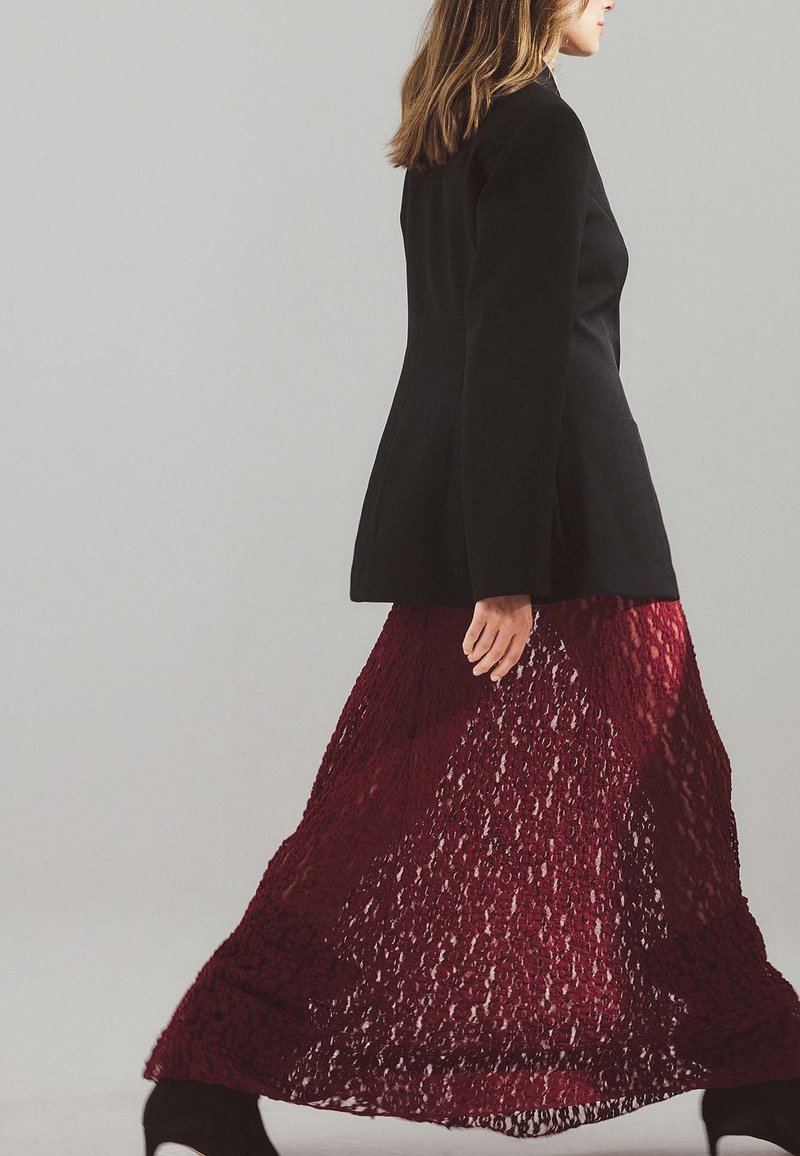 Frau, die in einem schwarzen Blazer, einem transparenten dunkelroten Spitzenrock und schwarzen, hohen Stiefeletten vor einem schlichten, hellen Hintergrund geht.