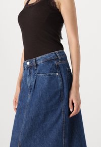 Denimihame keskisinisena; sisaldab kõrge vöökohaga, esipaneeliga nööpidest sulgemise ja küljetaskute funktsioone. Kangas on sileda tekstuuriga ja kergelt pleekinud.