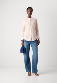 Camicia a maniche lunghe rosa chiaro, jeans a gamba larga blu, tiene in mano una piccola borsa blu, con sandali con tacco alti marroni, su uno sfondo neutro.