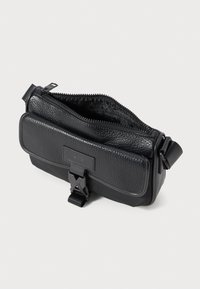 Melnā Armani Exchange pleca soma ar teksturētu priekšējo kabatu, ar rāvējslēdzēju galveno nodalījumu un regulējamu siksnu uz balta fona.