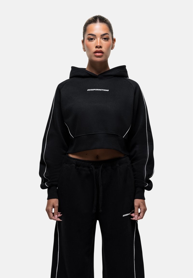 Haut de hoodie noir court avec un cordon de serrage, agrémenté de passepoils blancs et du texte « GOODFORMNOTHING ». Associé à un pantalon de jogging noir assorti.