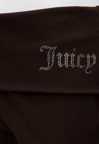 Σκούρο καφέ ύφασμα με τη λέξη "Juicy" γραμμένη με μικρές, λαμπερές στρας σε καλλιγραφική γραμματοσειρά.