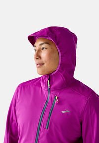 Fuchsia capuchonjack, gemaakt van lichte, rekbare stof. Voorzien van een rits aan de voorkant met neongele trekstangen en een logo op de linkerborst.
