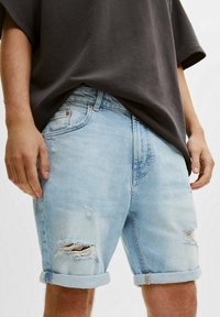Pantalones cortos de denim desgastado de color azul claro con los dobladillos doblados, usados por una persona con una camiseta de gran tamaño en gris oscuro, con las manos relajadas a los lados.